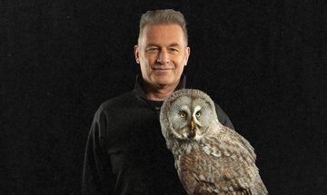 A close-up of Chris Packham who is wearing a black jumper. He is holding an owl that is staring forwards.