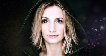 Cara Dillion, a very pale white woman with blonde hair, looking straight forward with very wide eyes and a neutral expression dressed in a hood with sparkles on the dark background behind her