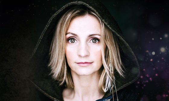 Cara Dillion, a very pale white woman with blonde hair, looking straight forward with very wide eyes and a neutral expression dressed in a hood with sparkles on the dark background behind her