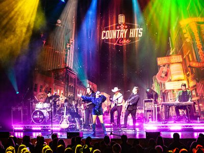 A colourfully lit stage with the backdrop of the Tennessee strip. There are six people on stage, three men playing guitar, one man on drums, one man on keyboard, and a woman singing passionately into a microphone with her other arm out stretched