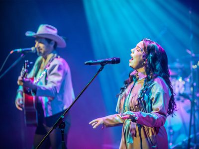 A man playing guitar and singing into a microphone in a white cowboy hat and matching jacket, next to a woman in a tan leather outfit who's also singing
