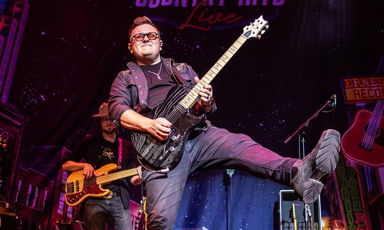 A man with short quiffed hair and glasses playing guitar on stage and kicking out his leg