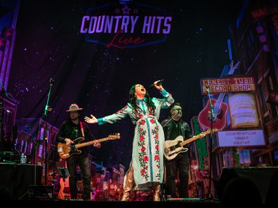 A woman in a long white and green dress with red flowers singing passionately into a microphone with two guitarists behind her