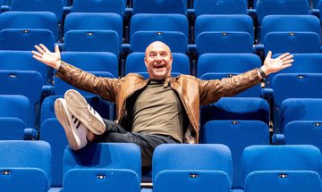 Chris Pizzey a bald white man in a brown leather jacket sat in the centre of blue seats with his arms out wide and feet up on the row of seats in front of him