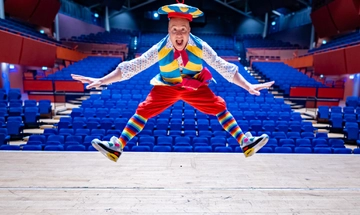 Chris Pizzey white man dressed in a colourful outfit mainly consisting of red, white, blue, and yellow. He is mid jump on The Anvil stage with his hat partially off his head