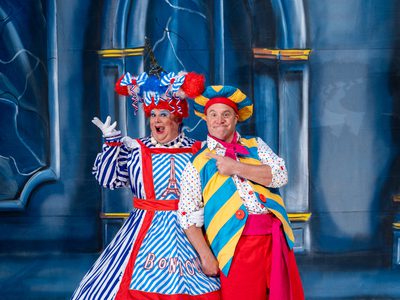 Richard J Hunt a white man dressed in drag. He is wearing a red, white, and blue puffy wig with an Eiffel Tower on top, and a large stripey blue and white dress with red accents and a matching apron that also has an Eiffel Tower and reds 'Bonjour'. He is stood smiling and posing with one arm in the air next to Chris Pizzey, a white man in red trousers with a long pink fabric belt, white shirt with multicoloured polka dots, a pink neck scarf, over sized yellow and blue stripped waist coast with pink buttons and a matching oversized beret