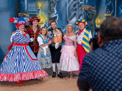 The Beauty and the Beast cast stood together in front of a castle background smiling and while a photographer takes their photo