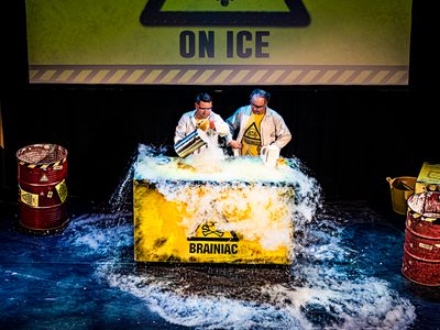 Two male scientists in lab coats pour dry ice over a large yellow box which reads Brainiac
