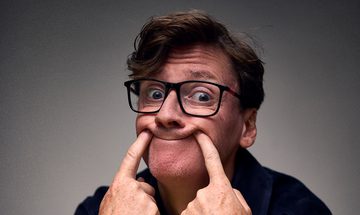 Ed Byrne, a white man with short quiffed brown hair and glasses, poking both of his index fingers into the corners of his mouth making an odd smile
