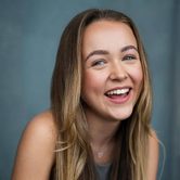 Ellie Dadd a young white blonde woman with long straight hair leaning forward in a grey vest top and a silver necklace and nose stud smiling and laughing
