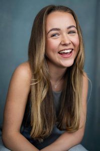 Ellie Dadd a young white blonde woman with long straight hair leaning forward in a grey vest top and a silver necklace and nose stud smiling and laughing