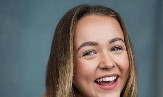 Ellie Dadd a young white blonde woman with long straight hair leaning forward in a grey vest top and a silver necklace and nose stud smiling and laughing
