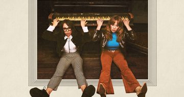 Flo and Joan sit on the floor in front of a piano, with their backs resting against the piano they reach up over and behind their heads and play the piano behind them