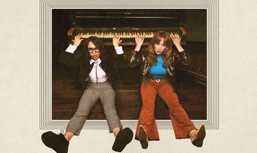 Flo and Joan sit on the floor in front of a piano, with their backs resting against the piano they reach up over and behind their heads and play the piano behind them
