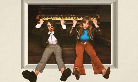 Flo and Joan sit on the floor in front of a piano, with their backs resting against the piano they reach up over and behind their heads and play the piano behind them