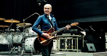Francis Rossi on stage in a blue jacket, white shirt, and black tie holding a guitar surrounded by instruments including a drum set and keyboard