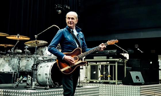 Francis Rossi on stage in a blue jacket, white shirt, and black tie holding a guitar surrounded by instruments including a drum set and keyboard