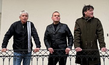 Three older men in black jackets leaning against a metal railing