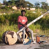Gasper Nali, a young black man in casual clothes, sat outside on a drum holding his babatoni, a very large instrument that looks similar to a guitar but with only one string. It has a round body, the same size as the drum he's sat on, and a long thin neck that looks like an old fence post