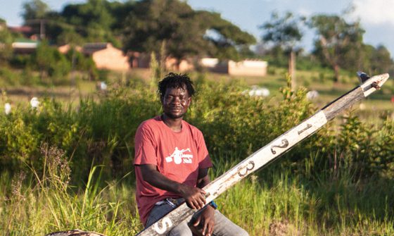 Gasper Nali, a young black man in casual clothes, sat outside on a drum holding his babatoni, a very large instrument that looks similar to a guitar but with only one string. It has a round body, the same size as the drum he's sat on, and a long thin neck that looks like an old fence post