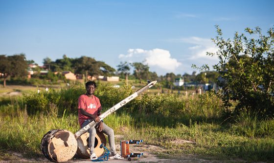 Gasper Nali, a young black man in casual clothes, sat outside on a drum holding his babatoni, a very large instrument that looks similar to a guitar but with only one string. It has a round body, the same size as the drum he's sat on, and a long thin neck that looks like an old fence post