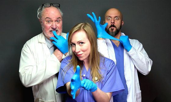 Three people, two men and a woman, all stood in doctors uniforms playing with blue rubber gloves