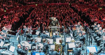 The Grimethorpe Colliery Band performing on stage bathed in white light with the audience behind in red light