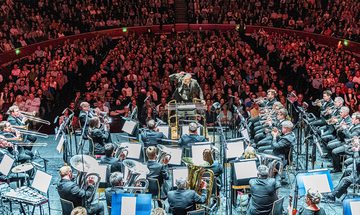 The Grimethorpe Colliery Band performing on stage bathed in white light with the audience behind in red light