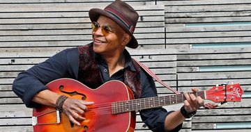 Guy Davis, an older black man in a round orange sunglasses, black shirt, brown waist coat, and hat, playing guitar