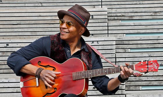 Guy Davis, an older black man in a round orange sunglasses, black shirt, brown waist coat, and hat, playing guitar