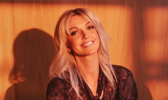 a blonde woman smiles at the camera, she wears jeans a a patterned shirt