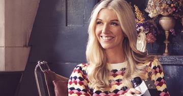 A blonde woman smiling in a chair in a multicoloured jumper holding an old camera with dry flowers behind her