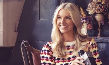 A blonde woman smiling in a chair in a multicoloured jumper holding an old camera with dry flowers behind her