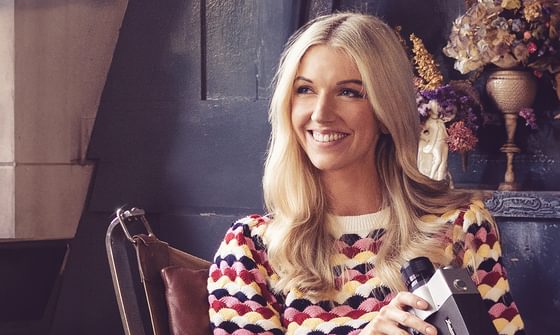 A blonde woman smiling in a chair in a multicoloured jumper holding an old camera with dry flowers behind her