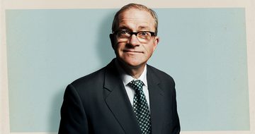Harry Enfield in a suit, tie, and glasses smiling on a blue and cream background