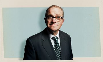 Harry Enfield in a suit, tie, and glasses smiling on a blue and cream background