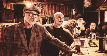 A sepia tone image of five older men stood at a bar with drinks. The man closest to the camera is staring at while everyone else isn't making any eye contact