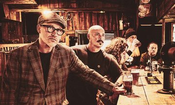 A sepia tone image of five older men stood at a bar with drinks. The man closest to the camera is staring at while everyone else isn't making any eye contact