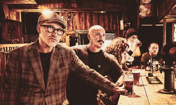 A sepia tone image of five older men stood at a bar with drinks. The man closest to the camera is staring at while everyone else isn't making any eye contact