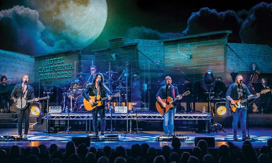 Four men playing guitars stand in a line across the front of the stage. Behind them are other musicians and a projection of a moon and a sign that reads Hotel California Tour 2026