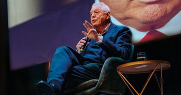 John Simpson sat on stage speaking into a microphone with a large picture behind him where you can make out the bottom of Donald Trump's face