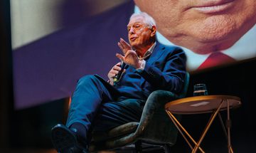 John Simpson sat on stage speaking into a microphone with a large picture behind him where you can make out the bottom of Donald Trump's face