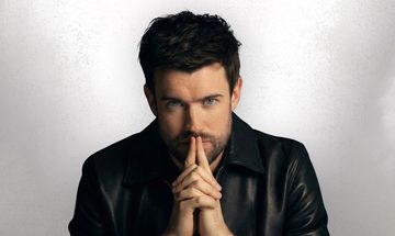 Jack Whitehall, a young white man with dark short scruffy hair and a stubbly beard in a leather jacket. His hands are clasped together with his index fingers extended, he is leaning on those fingers with them covering his mouth slightly