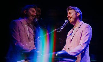 James B Partridge stands at a keyboard, he smiles as he sings into a microphone which is held in a stand. He's wearing a light pink suit and there's an effect on the photo which reflects his image with a rainbow lens flair over it