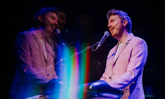James B Partridge stands at a keyboard, he smiles as he sings into a microphone which is held in a stand. He's wearing a light pink suit and there's an effect on the photo which reflects his image with a rainbow lens flair over it
