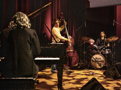 Three performers play on stage. You can see the back of the pianist and then facing them is the cellists and to the right of him is the drummer