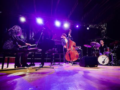 A cellists stands in the centre of a blue-lit stage, to the left is the pianist and to the right is the dummer
