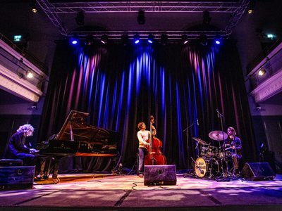 A cellists stands in the centre of a blue-lit stage, to the left is the pianist and to the right is the dummer