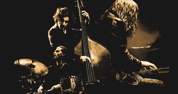 A sepia image of a drummer, a pianist and a cellists all performing