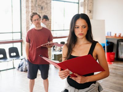 Jim Fish and Amy Tara rehearse a scene with serious expressions, each holding a script, while another person stands mostly behind Jim and is only partially visible.